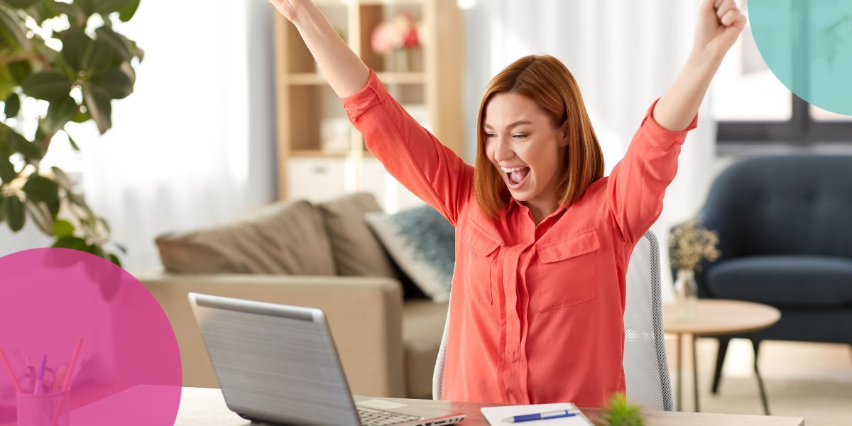 Woman sat at desk with her arms in the air whilst smiling at a laptop screen